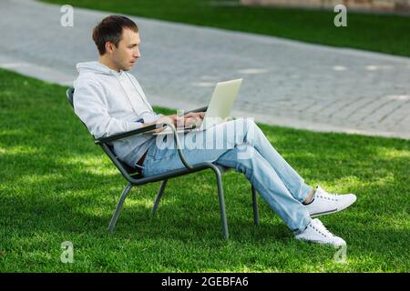 Blogger Mann arbeitet am Laptop im öffentlichen Park, verarbeitet Videos für Social-Media-Inhalte, sitzt im Stuhl Stockfoto