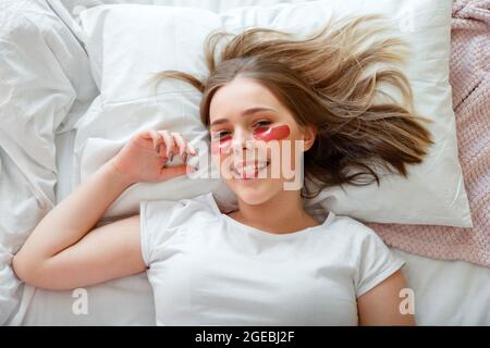 Junge Frau, die morgens Spaß hat und Zunge zeigt, während sie im Bett liegt. Teenager Mädchen haben Morgen Hautpflege Routine. Teenager-Mädchen mit Hydrogel-Auge Stockfoto