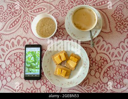 Auf einem mit rot-weißer Lacy-Tischdecke überzogenen Tisch befinden sich ein Mobiltelefon, eine Schüssel mit braunem Zucker, ein Teller mit Keksen und Käse sowie eine Cu Stockfoto