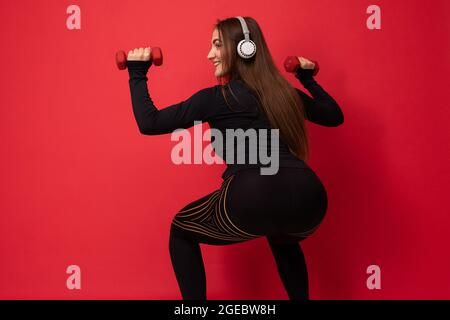 Schöne glücklich lächelnde junge Brunet Frau trägt schwarze Sportkleidung isoliert auf rotem Hintergrund Wand tun Fitness Squating mit Hanteln tragen Stockfoto