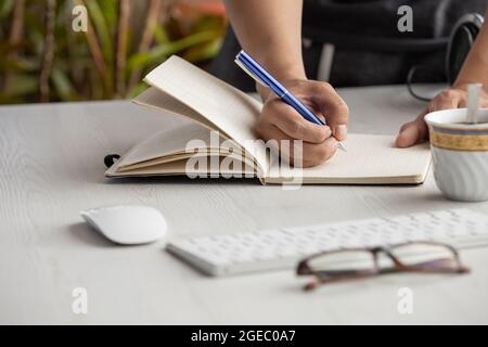 Nahaufnahme der Hand einer Person, die mit einem Stift auf einem Notebook schreibt, Arbeitsplatz mit Brille, Computermaus und Tastatur. Bürobedarf Stockfoto