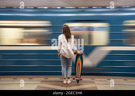 Mädchen mit Longboard steht auf U-Bahn-Plattform mit verschwommenen blauen Zug auf dem Hintergrund Stockfoto