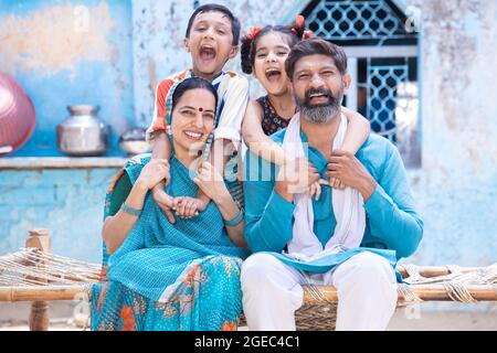 Fröhliche ländliche indische Familie auf dem traditionellen Bett im Dorf zu Hause sitzen, aufgeregt kleine Kinder Spaß mit den Eltern. Tochter und Sohn umarmen Vater und Stockfoto