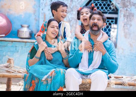 Fröhliche ländliche indische Familie auf dem traditionellen Bett im Dorf zu Hause sitzen, aufgeregt kleine Kinder Spaß mit den Eltern. Tochter und Sohn umarmen Vater und Stockfoto