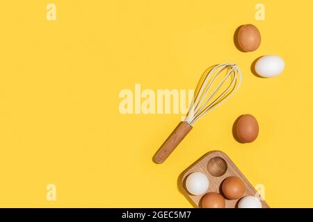 Eier in Holzkiste und Schneebesen auf gelbem Hintergrund. Flach liegend Draufsicht Kopierbereich. Stockfoto