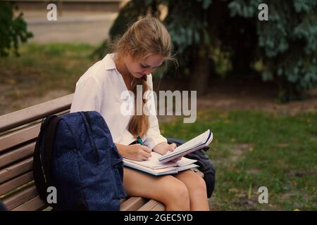 Eine junge Universitätsstudentin, die auf einer Bank sitzt, schreibt eine Zusammenfassung in einem Notizbuch aus einem Lehrbuch. Girl schreibt eine Zusammenfassung in einem Notizbuch aus einem textb Stockfoto