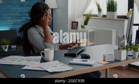 Müde verzweifelte schwarze Student leiden Kopfschmerzen sitzen am Schreibtisch im Wohnzimmer auf der Suche nach medizinischen Behandlung mit Computer. Besorgt krank verärgert junge Frau mit Migräneschmerzen während Lockdown Stockfoto