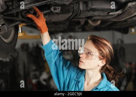 Die junge Cavsian-Automechanikerin an einer Autodienststelle steht unter einem erhöhten Auto und inspiziert das Chassis. Bekleidet mit einem blauen Overall. Stockfoto