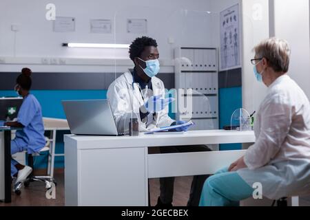afroamerikanischer Arzt, der am Schreibtisch sitzt und mit einer alten Frau über die medizinische Behandlung von Krankheiten spricht. Schwarzer Sanitäter, der während einer Coronavirus-Pandemie ältere Patienten mit Gesichtsmasken konsultiert Stockfoto