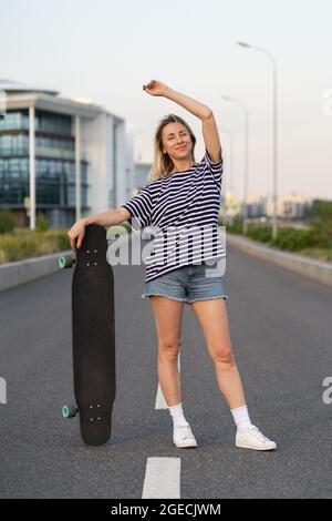 Erwachsene Frauen laufen gerne auf Longboard auf einer leeren Stadtstraße. Glückliche fröhliche Frau hält Skateboard Stockfoto