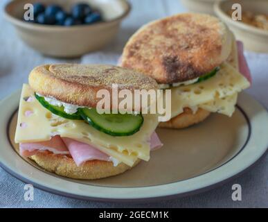 Frühstückssandwiches mit getoasteten englischen Muffins und gekrönt mit Schinken, deutschem Käse, Frischkäse und Gurken, serviert mit frischen Blaubeeren Stockfoto