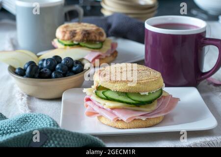 Gesundes Frühstückssandwich mit geröstetem englischen Muffin, gekrönt mit Schinken, Käse und Gurken, serviert mit Obst und einer Tasse Tee Stockfoto