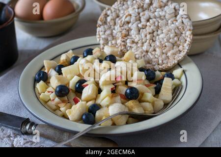 Gesundes glutenhaltiges und vegetarisches Frühstück mit einem frischen Obstsalat und einem aufgeblasenen Reiskuchen und gekochten Eiern, die zu Hause auf einem Tisch serviert werden Stockfoto