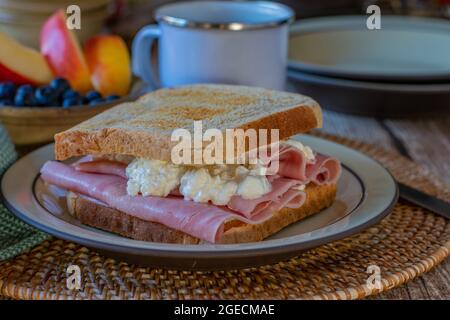 Getoastetes Frühstückssandwich mit Vollkorn-Toast, dünn geschnittenem Schinken und Quark. Serviert mit einer Tasse Kaffee und einer Schüssel mit Früchten Stockfoto