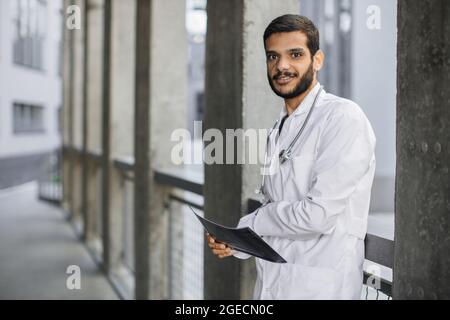 Waist up Outdoor-Porträt eines jungen, gutaussehenden, selbstbewussten männlichen arabisch-indischen Arztes, der die Kamera anschaut, den Röntgenscan des Patienten hält und in der Nähe des modernen H posiert Stockfoto
