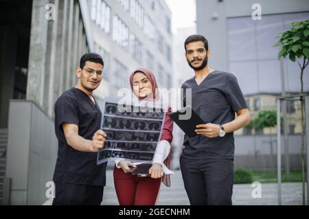Drei Ärzte, eine Frau im Hijab und zwei Männer in medizinischer Kleidung, diskutieren über die Röntgentomographie des Patienten und gehen vor dem Hintergrund der Moderne nach draußen Stockfoto