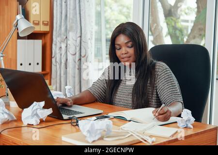 Weibliche Marketingmanagerin beantwortet E-Mails und Nachrichten auf einem Laptop und notiert sich im Planer, wenn sie am unordentlichen Schreibtisch arbeitet Stockfoto