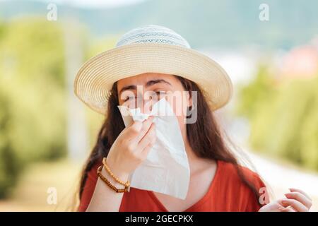 Eine Frau in einem Strohhut bläst ihre Nase in ein Taschentuch.Outdoor. Konzept von Allergie und Virus. Stockfoto