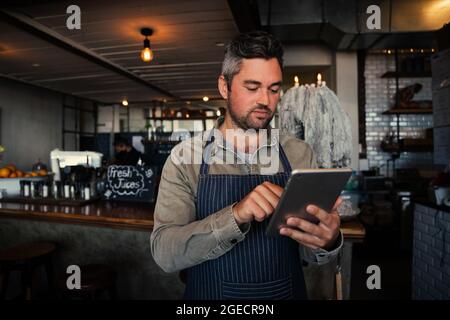 Kaukasischer Kellner scrollt durch das Online-Menü und steht in einem trendigen Café, wo er Bestellungen vorbereitet. Stockfoto