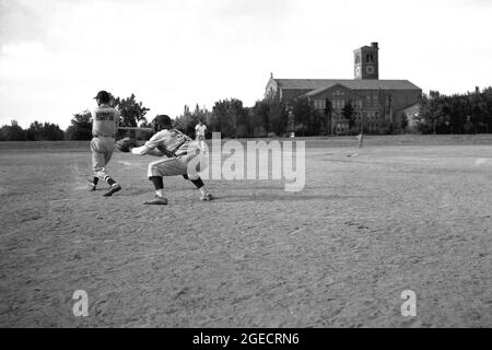 Historische 1950er Jahre, draußen auf einem staubigen Gebiet mit offenem Boden oder Sandplatz, lokale Bauarbeiter spielen ein Baseballspiel, Mid-West, USA. Hier sehen wir einen Schläger schwingen die Fledermaus, mit Fänger hinter, beide in voller Baseballausrüstung. Stockfoto