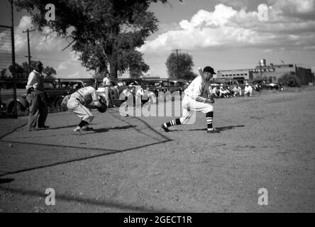 Die 50er Jahre, historisch, draußen auf einem staubigen Gebiet mit offenem Boden oder einem Ballfeld, neben einer Fabrik und mit Autos der damaligen Zeit geparkt, spielen lokale Bauarbeiter in voller Ausrüstung ein organisiertes, wettbewerbsorientiertes Baseballspiel, Mid-West, USA. Hier sehen wir einen Teig, einen Fänger und einen Hausschmier. Stockfoto