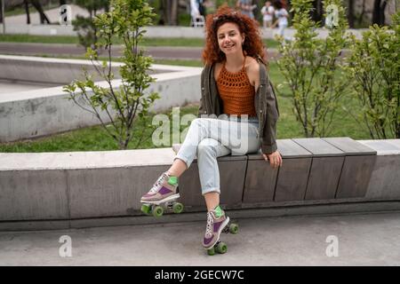 Fröhlicher und lockiger Rollerskater, der auf der Bank im Skatepark vor der Kamera lächelt Stockfoto