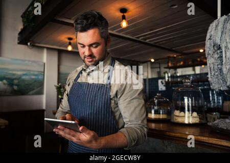 Hübscher kaukasischer Kellner, der auf dem digitalen ipad scrollt und sich beim Stehen im Café anmeldet Stockfoto