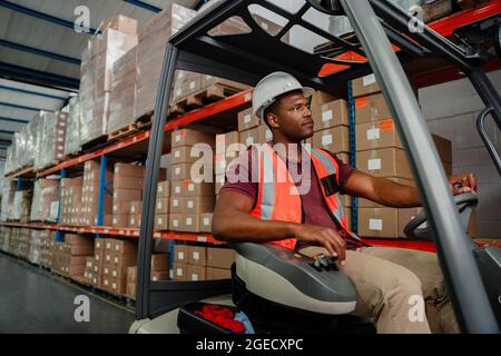Ethnischer Arbeiter, der im Gabelstapler um das Lager herum fährt und nach Paketen sucht, um die Lieferung zu versenden. Stockfoto
