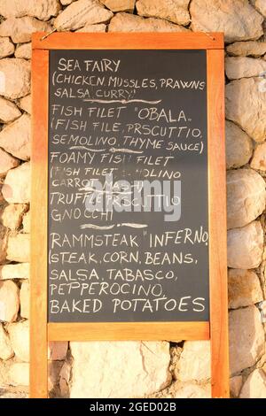 Schwarzes Tafelmenü, schwarze Farbe, Nachricht oder Werbung. Tafel steht in der Nähe des Restaurants an der Küste. Sonniger Tag. Stockfoto