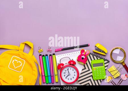 Zurück zur Schule, Bildungskonzept. Gelber Rucksack mit Schulbedarf - Notizbuch, Stifte, Lineal, Taschenrechner, Schere isoliert auf violettem Hintergrund Stockfoto