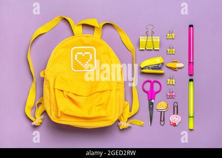 Zurück zur Schule, Bildungskonzept. Gelber Rucksack mit Schulbedarf - Notizbuch, Stifte, Lineal, Taschenrechner, Schere isoliert auf violettem Hintergrund Stockfoto