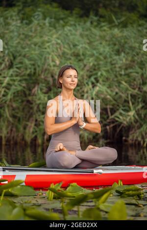 Ein junges kaukasisches, schönes, schlankes Mädchen, das Yoga macht, Pilates auf dem Paddle Board, SUP allein am Sommermorgen. Aktives Leben, Sport, Freizeitbeschäftigung Konzept Stockfoto