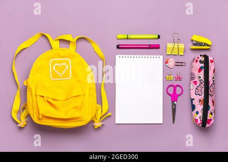 Zurück zur Schule, Bildungskonzept. Gelber Rucksack mit Schulbedarf - Notizbuch, Stifte, Lineal, Taschenrechner, Schere isoliert auf violettem Hintergrund Stockfoto
