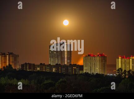 Tiefnachtsaufnahme eines Vollmondes über Wohnhäusern am Stadtrand. Eine helle Mondscheibe beleuchtet hohe Gebäude mit Licht in den Fenstern Stockfoto