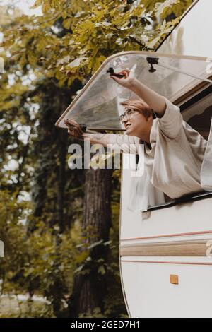 Junge Frau öffnet das Fenster des Campingwagens am Morgen im Herbstwald. Stockfoto