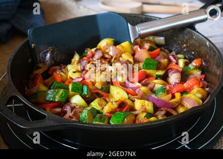 Gebratenes Gemüse in einer rustikalen gusseisernen Pfanne serviert. Mit roten Zwiebeln, Zucchini, roten Paprika und Knoblauch. Serviert auf einem Holztisch Stockfoto