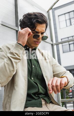 Junger Mann, der eine stilvolle Sonnenbrille anpasst, während er auf die Armbanduhr schaut Stockfoto