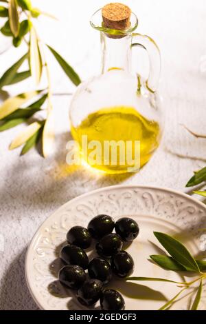 Ein Glas mit nativem Olivenöl, strukturierte Platte mit schwarzen Oliven und einem Zweig von Olivenbaum Stockfoto