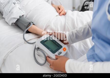 Beschnittene Ansicht einer afroamerikanischen Krankenschwester mit digitalem Tonometer in der Nähe des Patienten auf dem Krankenhausbett Stockfoto