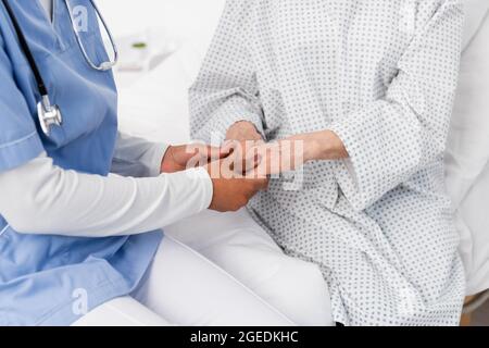 Beschnittene Ansicht einer afroamerikanischen Krankenschwester, die die Hände eines älteren Patienten im Kleid hält Stockfoto