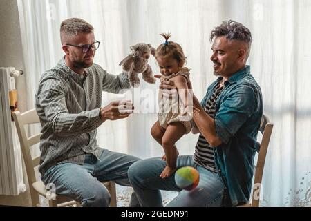 Schwule Männer mit adoptierten Mädchen zu Hause - zwei gutaussehende Väter spielen mit ihrer Tochter - LGBTQ+ Familie zu Hause - Diversity-Konzept und LGBT Stockfoto