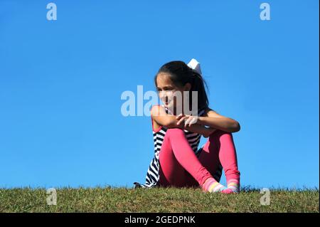 Kleines Mädchen sitzt und allein auf einem Hügel. Sie schaut seitwärts und ist in Gedanken verloren. Ein leuchtender blauer Himmel umgibt sie. Stockfoto