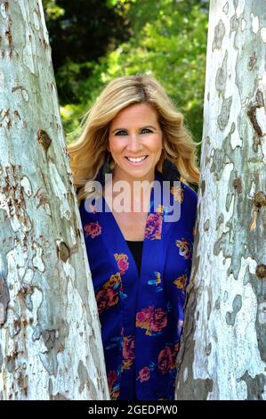 Die junge Frau lehnt sich an den Stamm eines Baumes und lächelt. Sie trägt eine Jacke mit blauem Druck und hat blonde Haare. Stockfoto