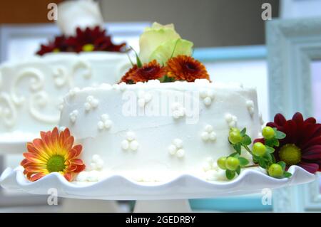Weiß vereister Hochzeitstorte ist mit Herbstblumen in Burgund und Orange verziert. Es setzt sich auf einen weißen, gläsernen Kuchenteller. Stockfoto
