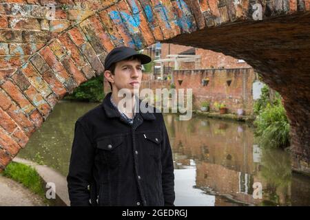 Ein junger Mann steht auf einem Kanalpfad in der Nähe einer Brücke. Er schaut den Kanal hinunter und wartet vielleicht auf jemanden. Stockfoto