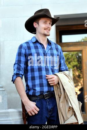Ein hübscher junger Mann, der Cowboyhut trägt und einen Mantel trägt, lächelt während einer westlichen Nachstellung das Publikum an. Stockfoto