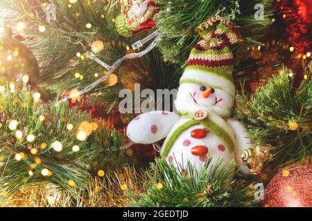 Lächelnder Schneemann aus Textil auf dem Weihnachtsbaum mit bunten, unscharfen Lichtern. Selektiver Fokus. Weihnachtskarte mit Dekorationen für den Silvesterabend Stockfoto