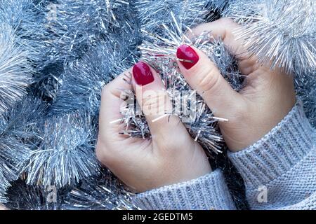 Weibliche Hände in einem grauen Strickpullover mit einer schönen glänzenden Maniküre - burgunderrote, dunkelrote, kirschfarbene Nägel auf dem Hintergrund einer silbernen Weihnachtsdose Stockfoto