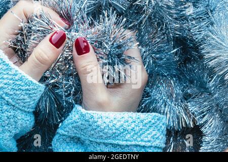 Weibliche Hände in einem blauen Strickpullover mit einer schönen glänzenden Maniküre - burgunderrote, dunkelrote, kirschfarbene Nägel auf dem Hintergrund einer silbernen Weihnachtsdose Stockfoto