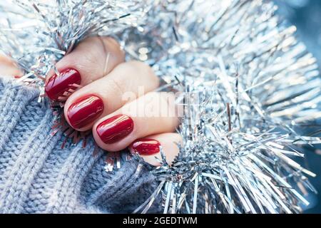 Weibliche Hände in einem grauen Strickpullover mit einer schönen glänzenden Maniküre - burgunderrote, dunkelrote, kirschfarbene Nägel auf dem Hintergrund einer silbernen Weihnachtsdose Stockfoto